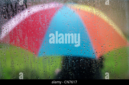 Gocce di pioggia su un vetro di finestra con una persona in possesso di un arcobaleno colorato ombrellone esterni Foto Stock