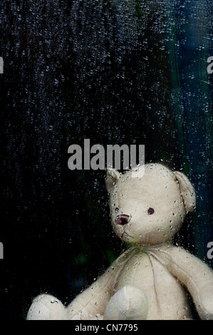 Triste orsacchiotto guardando attraverso un vetro ricoperto di gocce di pioggia. Still Life Foto Stock