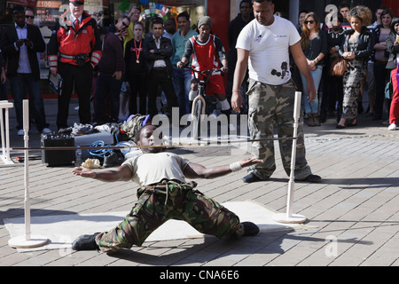 Gli artisti di strada del limbo danzando sotto una bassa la pole con un pubblico di persone che guardano in Leeds City Centre Yorkshire England Regno Unito Foto Stock