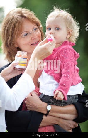 Carino bambina all'aperto in un parco essendo alimentato purea di mela da sua mamma e tenuto da sua nonna (SHALLOW DOF, il fuoco selettivo) Foto Stock