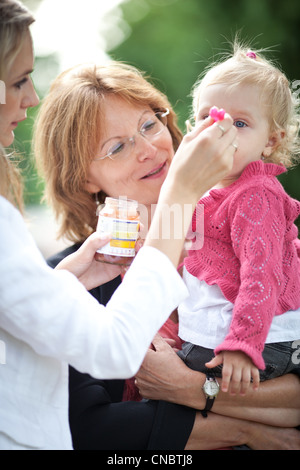 Carino bambina all'aperto in un parco essendo alimentato purea di mela da sua mamma e tenuto da sua nonna (SHALLOW DOF, il fuoco selettivo) Foto Stock