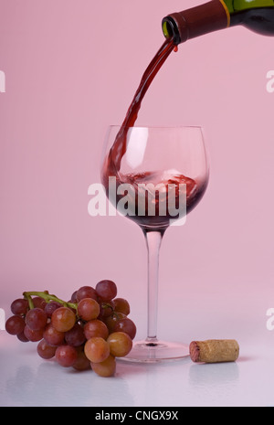 Vino rosso dal versamento di una bottiglia in un bicchiere di vino con uve a bacca rossa e un tappo di sughero accanto al vetro su una rosa di sfondo colorato. Foto Stock