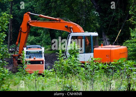Un teleobiettivo colpo di un retroescavatore e un piccolo caricatore a estremità anteriore insediamento folle in una radura in un campo incolto su un nuvoloso giorno. Foto Stock