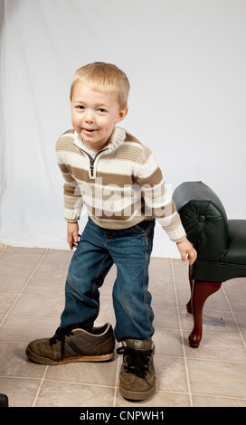 Carino il ragazzo biondo giocando e indossando i suoi padri scarpe, guardando la telecamera con una felice, giocoso sorriso Foto Stock