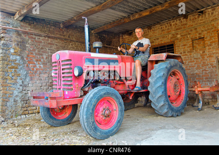 Burbero cercando conducente del trattore circum salariato agricolo non così felice supporto è il trattore in un granaio dopo una giornata nei campi Foto Stock