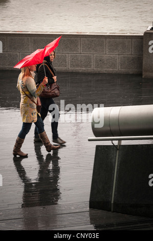 Più Londra Southbank due ragazze giovani donne condivisione spazzate dal vento rosso a piedi ombrello heavy rain riflessione terreno umido Fiume Tamigi Foto Stock