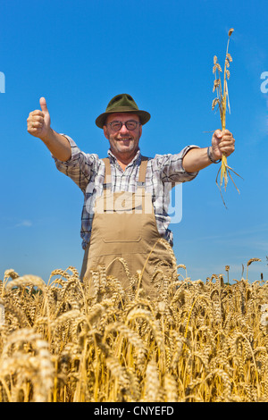 Pane di frumento, coltivati frumento (Triticum aestivum), agricoltore contently in piedi nella sua coppia campo di grano sollevando il pollice e spighe di grano, Germania Foto Stock