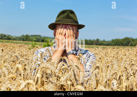 Frustrati agricoltore seduto nella sua coppia campo di grano tenendo la mano davanti al suo volto, Germania Foto Stock