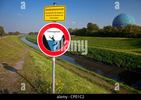 Segnale di avvertimento di drawning e aprire delle acque reflue canal Emscher con il contenitore di gas in background, in Germania, in Renania settentrionale-Vestfalia, la zona della Ruhr, Gelsenkirchen Foto Stock