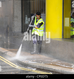 Uomo di scena di strada che lavora in giacca ad alta visibilità e indossare guanti occhiali & stivali wellington pressione acqua lavaggio marciapiede marciapiede Londra Inghilterra Regno Unito Foto Stock