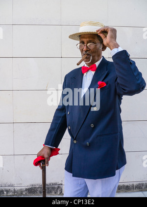 Un buio anziano gentiluomo cubano in un diportista più paglia hat, blu sport coat, red bow tie e un sigaro si erge da una parete a l'Avana. Foto Stock