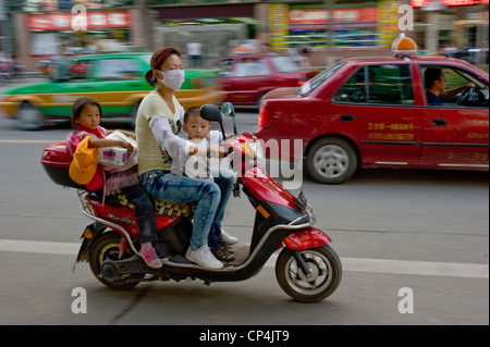 Un locale signora cinese a cavallo del suo scooter e 2 bambini non indossano caschi - con motion blur. Foto Stock