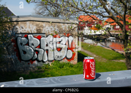 Rosso Coca Cola può in piedi su un corrimano grigio in background su un canale con albero in fiore e vantarti! Come graffiti su una soleggiata giornata di primavera Foto Stock