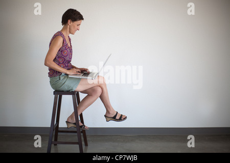 Donna di mezza età seduta su uno sgabello e utilizzando il suo laptop computer contro un muro grezzo. Foto Stock