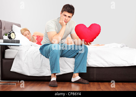 Giovane uomo in pensieri tenendo un cuore Cuscino sagomato mentre la sua fidanzata addormentarsi Foto Stock