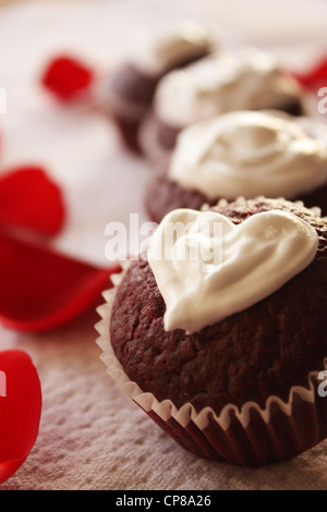 Il giorno di San Valentino a tema velluto rosso tortine con a forma di cuore di crema di formaggio topping. Petali di rosa sono visti in background Foto Stock