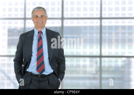 Sorridente di mezza età uomo di affari con le mani nelle tasche dei pantaloni. Formato verticale in piedi di fronte ad una grande finestra del moderno di Foto Stock