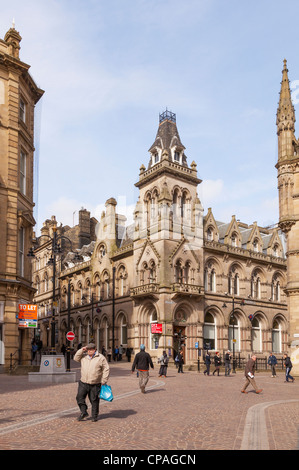 Un vecchio uomo in un tappo piatto cammina davanti a vecchi edifici in pietra arenaria a Bradford, West Yorkshire, Inghilterra, Foto Stock