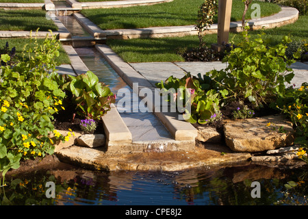 Un giardino con caratteristiche di acqua rigogliosa in pietra e bordi pavimentati prati prati piante marginali che piantano il Regno Unito Foto Stock