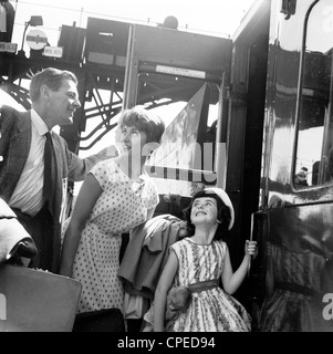1960s, storico, estate una famiglia eccitata che va in vacanza a bordo di un treno carrozza su una piattaforma ferroviaria, Inghilterra, Regno Unito. La donna indossa un abito spotted che era alla moda in questa epoca, mentre l'uomo è in forma vestito con una camicia e cravatta, con giacca, come era la norma in questo momento. La giovane figlia guarda in su ai suoi genitori nell'eccitamento. Foto Stock