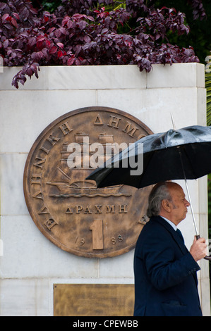 Un greco uomo a camminare con una lapide raffigurante una vecchia dracma moneta che è stato sostituito dall'euro nel 2002 fuori Athens City Hall Foto Stock