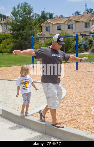 Portando come loro equilibrio su un cordolo, un padre gioca con la sua figlia in un Lake Forest, CA, parco. Nota attrezzature per parchi giochi. Foto Stock