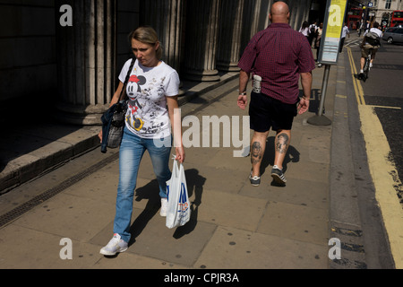 Un uomo con Laurel e Hardy tatuaggi passeggiate lungo un City of London street, passando una donna con Mickey Mouse sul suo T-shirt. Foto Stock