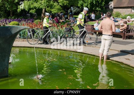 Due agenti di polizia con la loro bicicletta passano da un uomo nella fontana in una calda giornata estiva. Holland Park, Londra. Regno Unito. Foto Stock