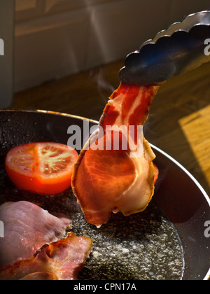 La pancetta la frittura SUN CUCINA PAN albero del sole illumina un rasher di organico back bacon essendo ruotata in una padella calda contenente pomodori Foto Stock