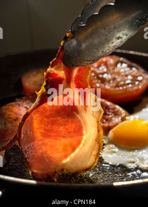 Uova pancetta pomodori la frittura in presenza di luce solare illuminazione di un rasher di organico back bacon essendo ruotata in una padella calda contenenti uova fritte e pomodori Foto Stock