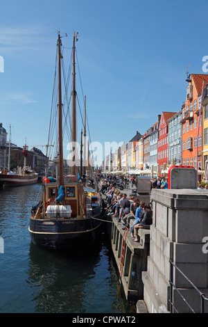 Vecchie imbarcazioni ormeggiate nel canale di Nyhavn a Copenaghen piena di rilassarsi e bere birra di persone su una soleggiata giornata di primavera Foto Stock