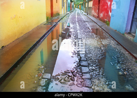 La riflessione in acqua di pioggia pozzanghere lungo la strada acciottolata in tradizionale vivacemente colorato street di Antigua Guatemala in estate. Foto Stock
