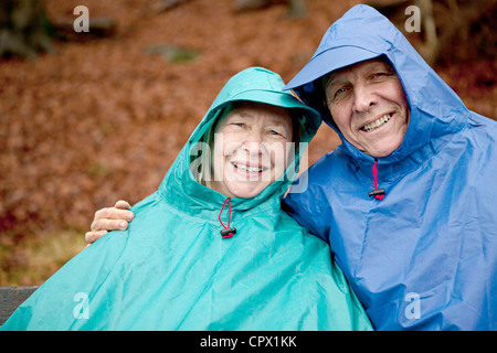 Ritratto di coppia senior seduto su una panchina nel parco indossando impermeabilizza Foto Stock