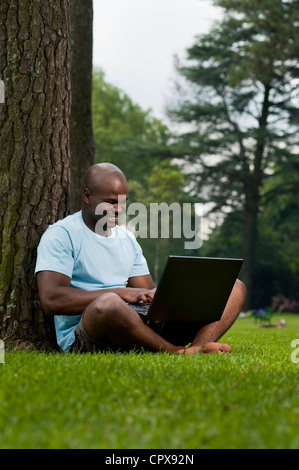 Giovane africano uomo seduto in un parco, al lavoro su un notebook Foto Stock