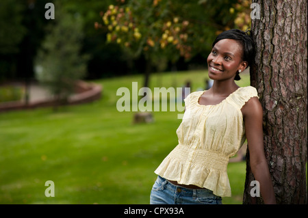 Giovane donna africana in piedi contro un albero in un parco, sorridente Foto Stock