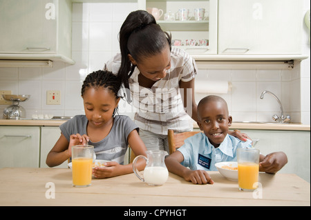 I giovani fratelli africani consumano la colazione insieme in una cucina, con la loro madre in background Foto Stock