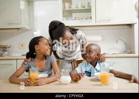 I giovani fratelli africani consumano la colazione insieme in una cucina, con la loro madre in background Foto Stock