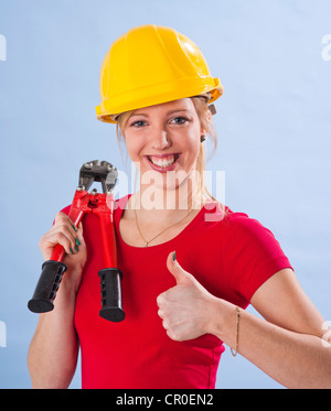 Giovane donna che indossa un giallo casco di sicurezza e tenendo un bullone coltello nella sua mano, rendendo un pollice in alto gesto Foto Stock