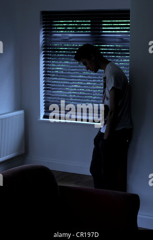 Giovane maschio in piedi in una stanza buia appoggiata contro il muro dietro di lui è una finestra cieca. Modello e proprietà (proprietà di fotografo) rilasciato. Foto Stock