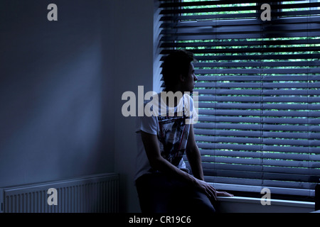 Giovane maschio seduto in una stanza buia guardando fuori attraverso una finestra cieca. Modello e proprietà (proprietà di fotografo) rilasciato. Foto Stock
