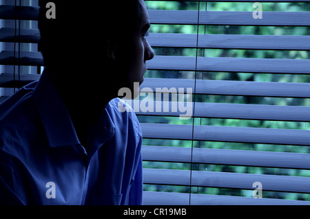 Giovane uomo seduto in una stanza buia guardando fuori attraverso una finestra cieca. Foto Stock