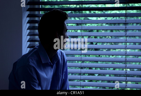 Giovane uomo seduto in una stanza buia guardando fuori attraverso una finestra cieca. Foto Stock