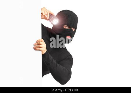 Un ladro con maschera di rapina tenendo una torcia dietro un pannello bianco isolato su sfondo bianco Foto Stock