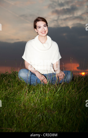 Sorridente giovane donna seduta in un prato, tramonto Foto Stock