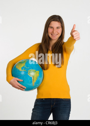 Ragazza adolescente holding globe e mostrando pollice in su contro uno sfondo bianco, sorridente, ritratto Foto Stock