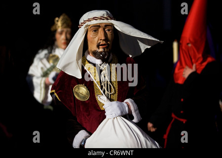Un uomo mascherato vestito come un personaggio biblico passeggiate passato un penitente durante una PASQUA SETTIMANA SANTA PROCESSIONE in Puente Genil, Spagna Foto Stock