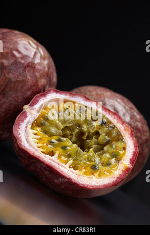 Due frutti della passione (Passiflora edulis), un frutto intero e uno tagliato a metà, al buio su un piano di vetro Foto Stock