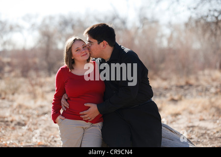 Carino felice incinta giovane coppia sposata all'aperto appoggiata contro un log. Foto Stock