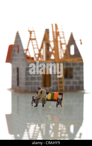 Figura in miniatura di una persona senza dimora seduta su una panchina di fronte a una casa non finita in costruzione, immagine simbolica di Foto Stock