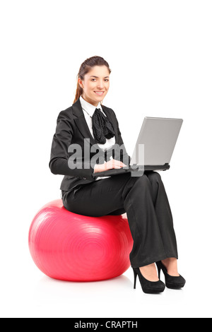 Donna sorridente seduto su una sfera di pilates e lavorando su un computer portatile isolata contro uno sfondo bianco Foto Stock